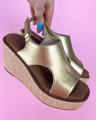 Gold wedge sandal with straw sole held by a hand against a pink background