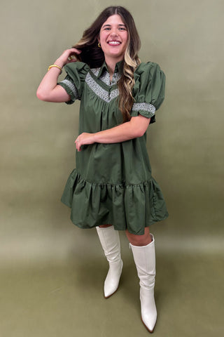 Woman wearing a green dress with white embroidery and white knee-high boots against a green background