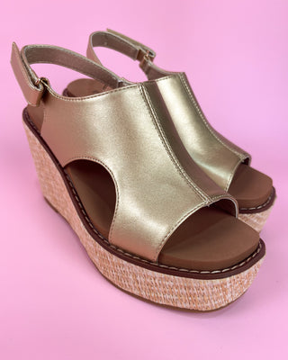 Gold wedge sandal with straw sole held by a hand against a pink background