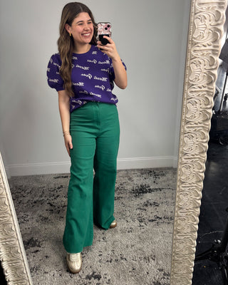 Woman taking a mirror selfie wearing a purple patterned top and green pants.