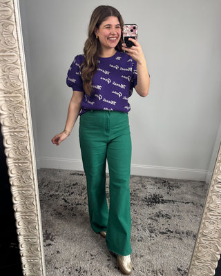 Woman taking a mirror selfie wearing a purple top with white text and green pants.