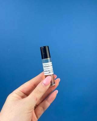 A hand with pink manicured nails holds a small clear glass roller bottle with a black cap and a white label against a solid blue background.
