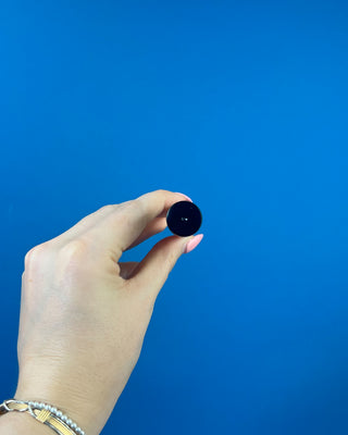 A hand with pink manicured nails holds a small clear glass roller bottle with a black cap and a white label against a solid blue background.