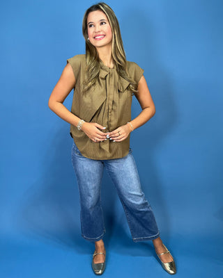 A woman modeling a sleeveless olive-brown ruffled blouse with blue cropped jeans and metallic flats, standing against a solid blue studio backdrop.