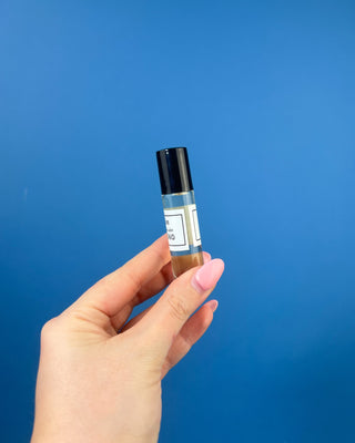 A small clear glass rollerball bottle with a white label and '100% Organic', shown on a wooden surface with a black cap beside it and a blue background.