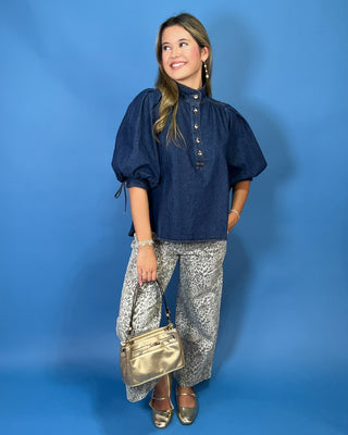 A woman modeling a dark blue chambray puff-sleeve blouse with button details, paired with wide-leg patterned gray pants, a metallic gold handbag, and gold flats