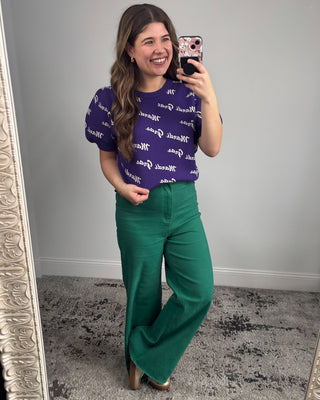Woman taking a mirror selfie wearing a purple sweater with white text and green pants.