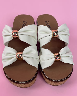Pair of sandals with large white bows and rose gold accents on a pink background