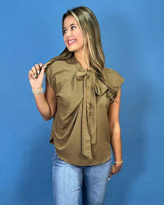 A woman modeling a sleeveless olive-brown top with a large front bow and ruffled shoulders, paired with blue jeans, against a solid blue studio background.