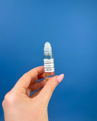 A hand with pink manicured nails holds a small clear glass roller bottle with a black cap and a white label against a solid blue background.