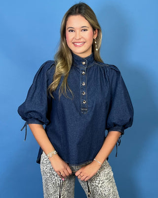 A woman modeling a dark blue denim puff-sleeve blouse with a buttoned high collar, paired with gray and white patterned pants