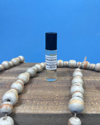 A hand with pink manicured nails holds a small clear glass roller bottle with a black cap and a white label against a solid blue background.