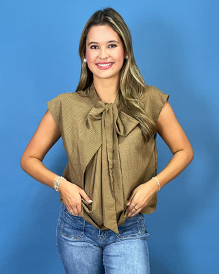 A woman modeling a sleeveless olive green top with a large front bow tie detail, paired with blue jeans, against a solid blue background.