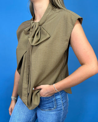 A woman wearing a sleeveless olive green top with a vertical stripe texture and a large bow detail at the collar, paired with blue jeans against a solid blue background.