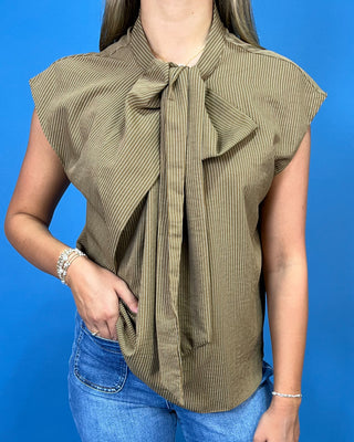 A woman wearing a sleeveless olive green top with vertical stripes and a large bow tie detail at the collar, paired with blue jeans, set against a solid blue background.