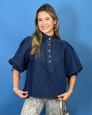 A woman modeling a dark indigo blue chambray puff-sleeve blouse with a buttoned high collar, paired with leopard-print pants