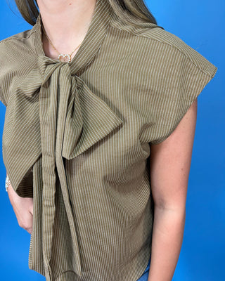 A close-up of a person wearing a short-sleeve olive green top with thin vertical stripes and a large bow tie at the collar, set against a solid blue background.