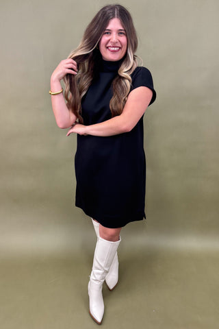 Woman in a black dress and white boots posing against a green background