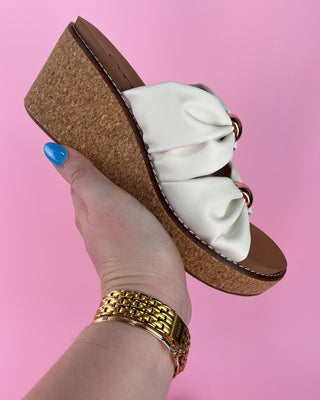 Brown wedge sandal with white bow held by a hand against a pink background