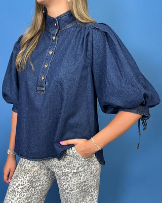 A woman wearing a dark blue denim blouse with puff sleeves, a buttoned high collar, and side tie details, paired with gray leopard print pants