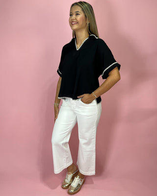 Woman wearing a black top with white trim and white pants against a pink background