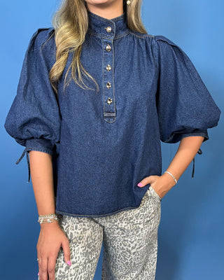 A woman wearing a dark blue denim blouse with puff sleeves, a high mock neck, and decorative gold-tone buttons down the front, paired with gray leopard print pants