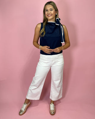 Woman wearing a navy sleeveless top with a white scarf and white pants on a pink background