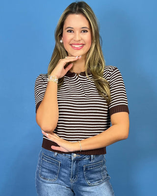  a brown and white horizontally striped short-sleeve knit top and blue jeans, posing against a solid blue studio background.
