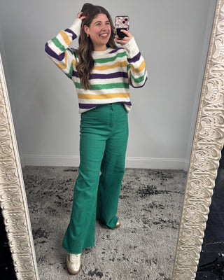 Person wearing a colorful striped sweater and green pants standing in front of a mirror.