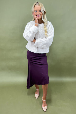 Woman wearing a white textured sweater and purple skirt against a green background
