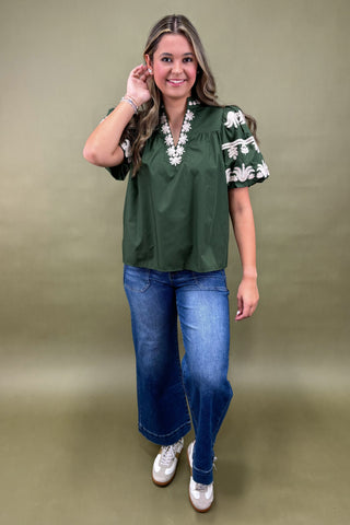 Woman wearing a green blouse with white floral patterns and blue jeans on a beige background