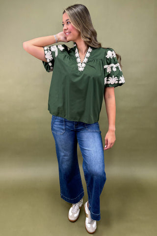 Woman wearing a green blouse with white embroidery and blue jeans on a beige background