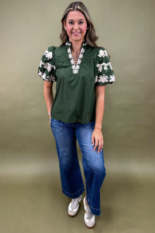 Woman wearing a green blouse with floral sleeves and blue jeans against a beige background