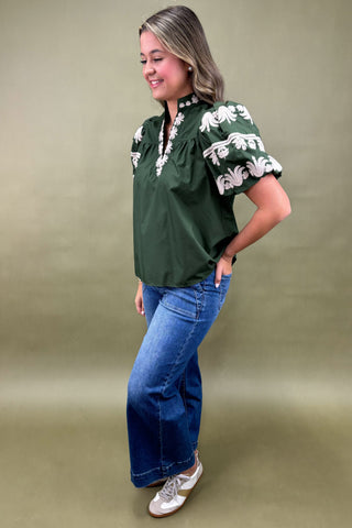 Woman wearing a green blouse with white floral embroidery and blue jeans on a beige background