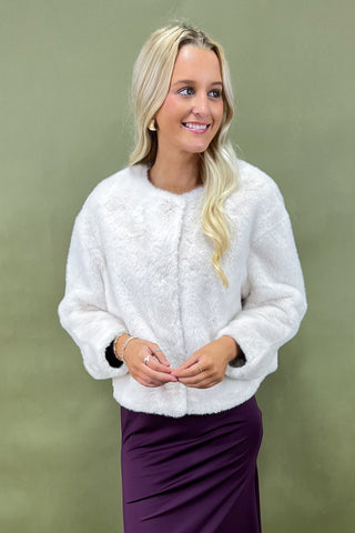Woman wearing a white textured sweater over a purple skirt against a green background