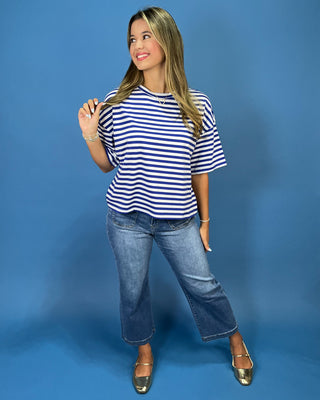 A woman modeling a navy and white horizontally striped short-sleeve t-shirt, paired with blue jeans and metallic gold flats, standing against a solid blue studio backdrop.