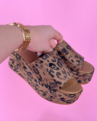Leopard print platform sandals held by a hand against a pink background