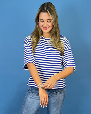 A woman wearing a blue and white horizontally striped short-sleeve top and blue jeans, standing against a solid blue studio background.