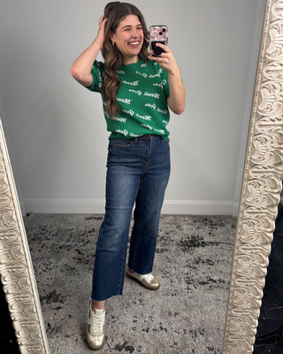 Woman taking a mirror selfie wearing a green patterned shirt and blue jeans.