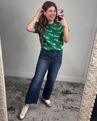 Woman taking a mirror selfie wearing a green t-shirt with text and blue jeans.