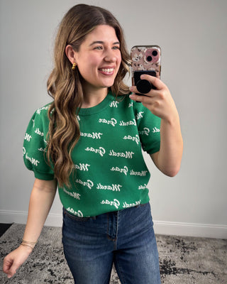 Woman taking a selfie wearing a green shirt with white text and blue jeans.