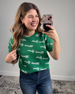 Woman taking a mirror selfie wearing a green shirt with white text and blue jeans.