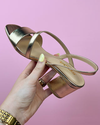 Gold sandal held by a hand with a pink background