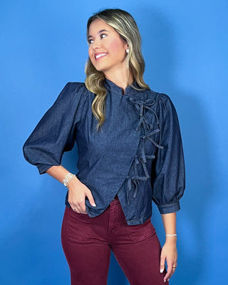 Dark wash denim tie-front top featuring a structured stand collar, puff three-quarter sleeves, asymmetrical front, and statement bow-tie closures.