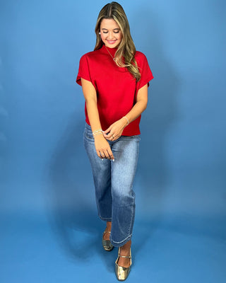A woman modeling a bright red short-sleeve mock neck top with blue jeans and metallic gold shoes, standing against a solid blue studio background.