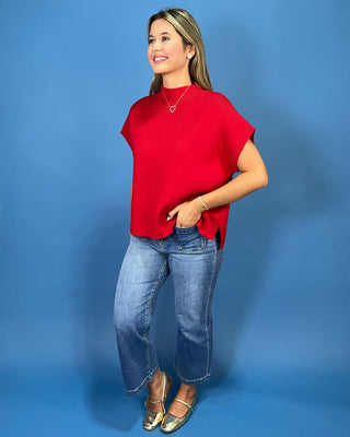 A woman modeling a bright red short-sleeve top with blue jeans and gold flat shoes, standing against a solid blue studio backdrop.