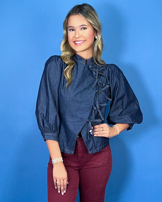 Dark wash denim tie-front top featuring a structured stand collar, puff three-quarter sleeves, asymmetrical front, and statement bow-tie closures.