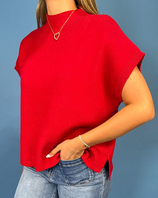 A woman wearing a bright red short-sleeve knit top with a mock neck and side slits, paired with blue jeans, against a solid blue background.