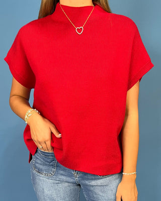 A woman wearing a bright red short-sleeve ribbed knit top with a mock neck, paired with blue jeans, against a solid blue background.