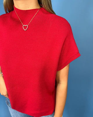 A person wearing a bright red short-sleeve ribbed knit top with a gold heart necklace, shown against a solid blue background.